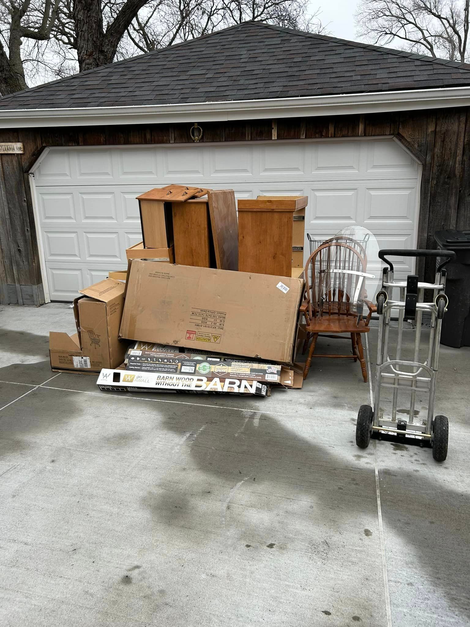 Pile of furniture and boxes in front of a garage. Includes a wooden chair, and a hand truck.