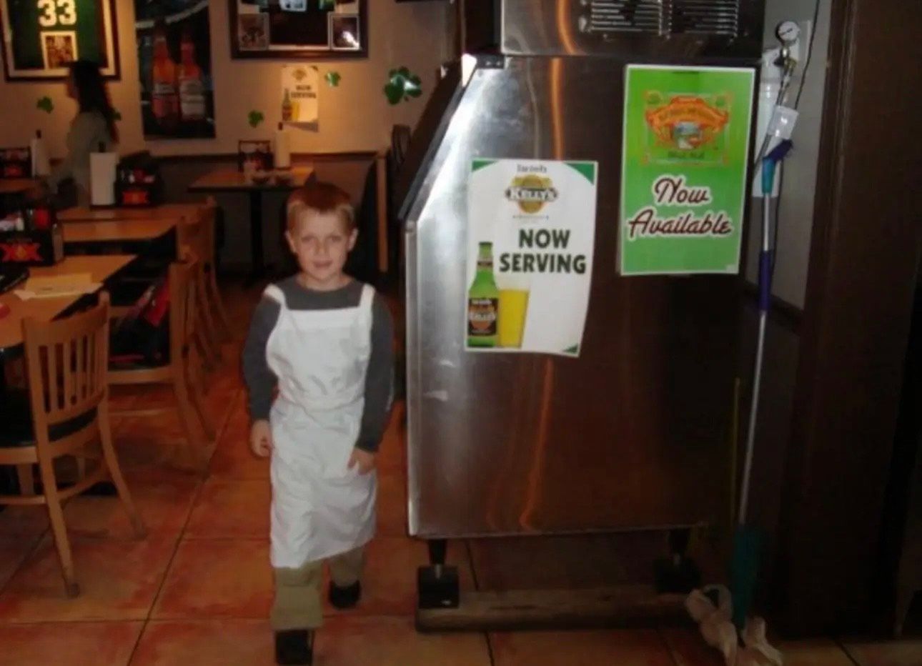 A young boy in an apron stands in front of a machine that says now serving