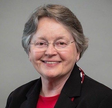 Woman with glasses smiles, wearing a black blazer over a red top, against a gray background.