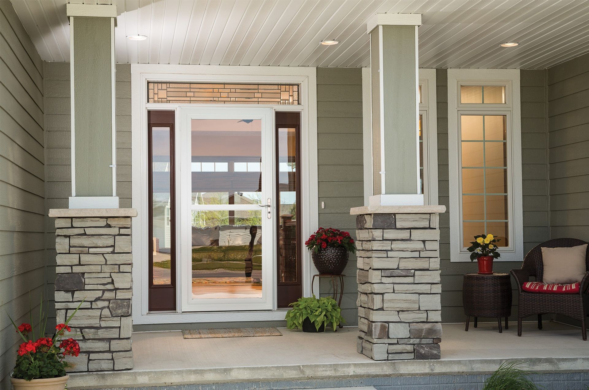 A front porch with stone-based pillars, a white storm door with sidelights, and a small sitting area with potted plants.