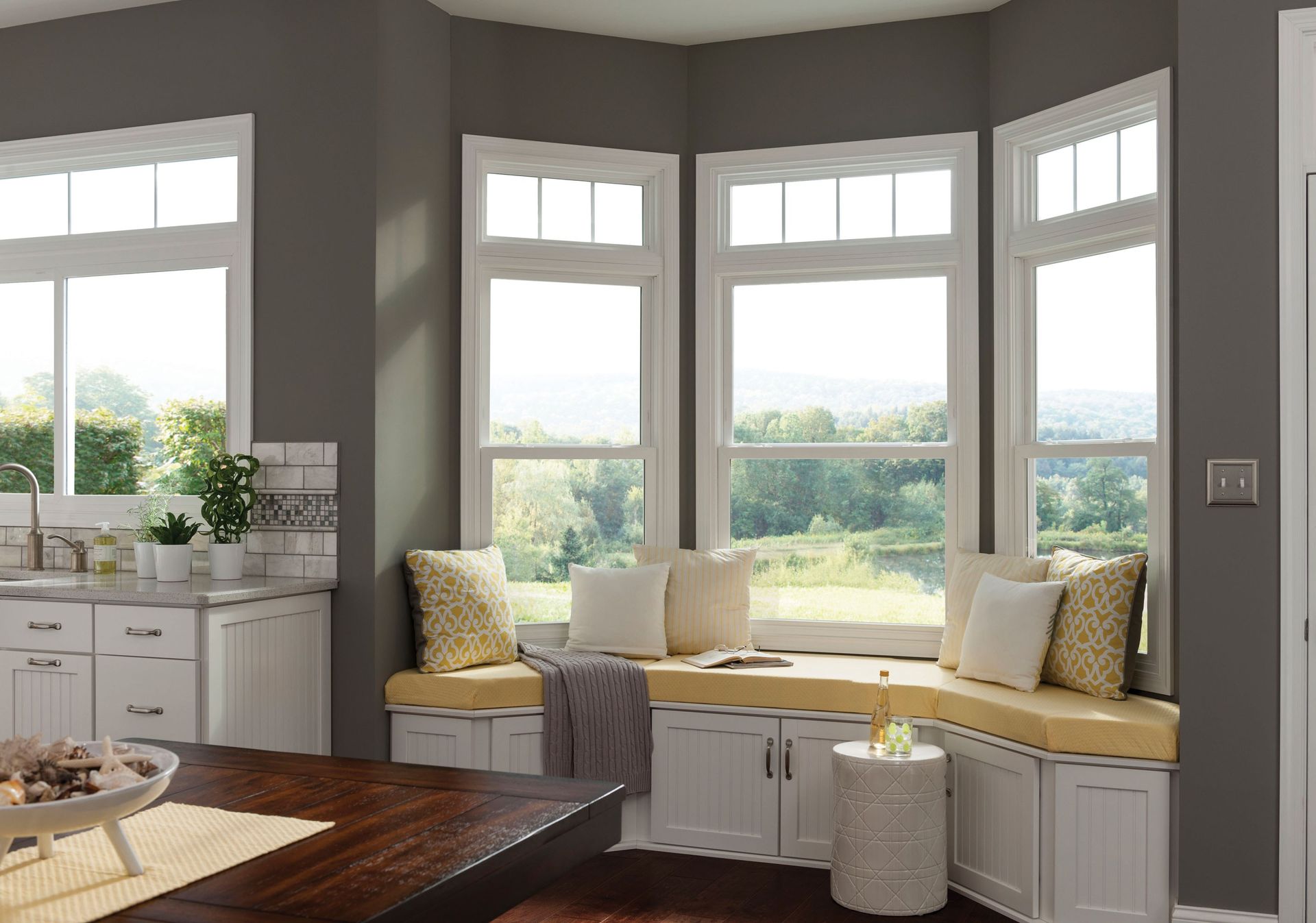 A kitchen bay window with built-in storage seating, yellow cushions, pillows, and white cabinets against dark gray walls.