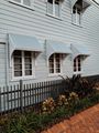 Light grey clapboard house with three windows featuring matching awnings, a dark picket fence, and a brick walkway.