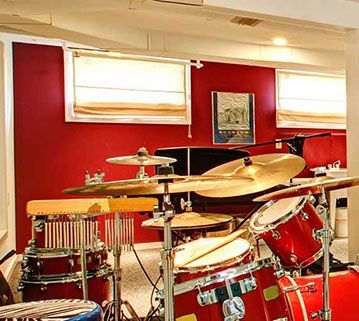 A red drum set stands in a basement room with red walls, two windows with blinds, and framed wall art.