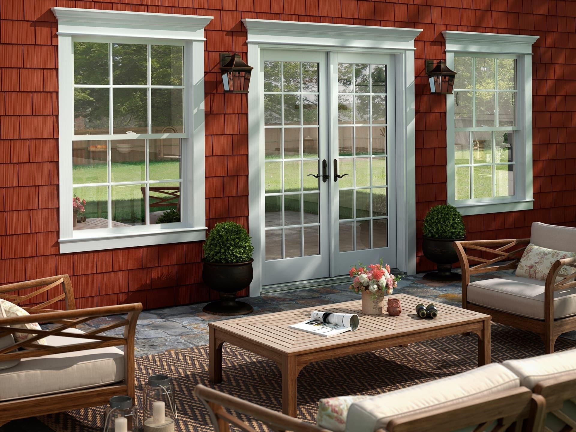 A patio area with a wooden coffee table and chairs in front of a red shingled house with white-framed doors and windows.