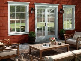 A patio area with a wooden coffee table and chairs in front of a red shingled house with white-framed doors and windows.