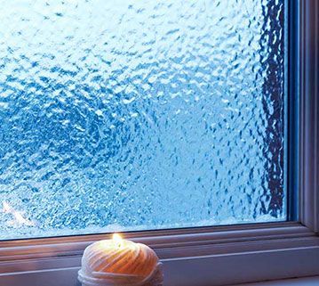 A lit white candle sits on a windowsill against a blue, textured, frosted glass window.