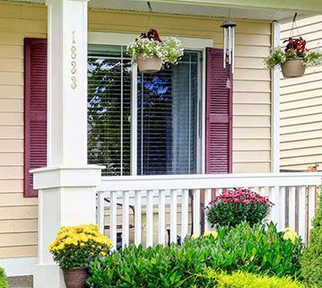 A front porch with yellow siding, maroon shutters, hanging flower baskets, and lush greenery in front of a window.