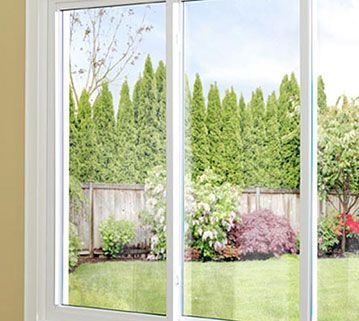 A white-framed sliding glass window looks out onto a green lawn, a wooden fence, shrubs, and a row of tall pine trees.