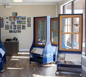 A showroom featuring multiple display stands with wood-framed windows, a desk, and a photo collage on the beige wall.