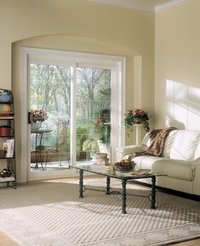 A bright living room with a sliding glass door opening to a garden, featuring a cream sofa, glass table, and area rug.
