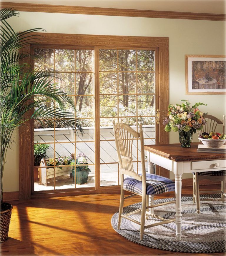 A wooden-framed sliding glass door overlooks a patio with potted plants, next to a dining set on a rug.