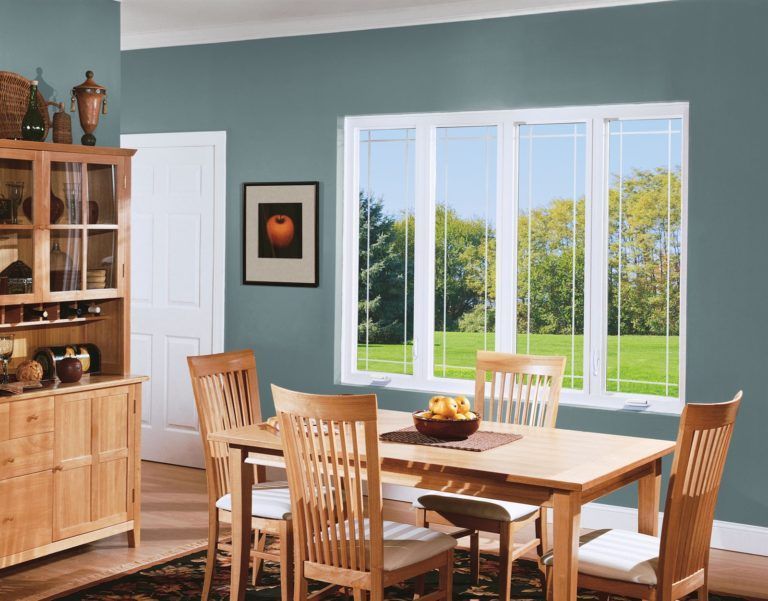 A wooden dining set with four chairs sits in front of a large, four-pane window against a blue-gray wall.