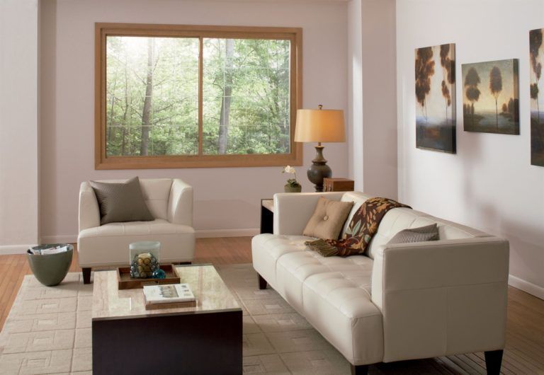 A bright living room features a beige sofa and armchair, a stone coffee table, and a large window looking onto a forest.