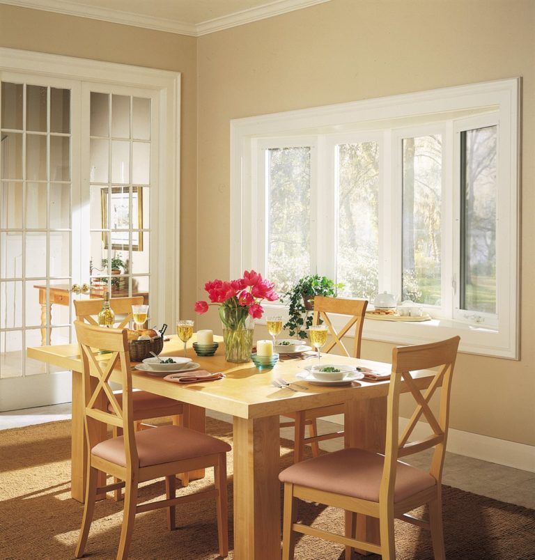 A light wood dining table with four chairs set in a bright room with a bay window and glass-paned doors.