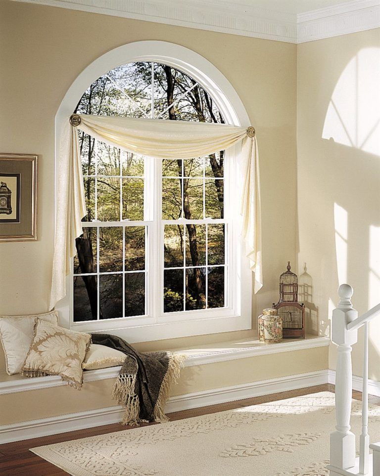 Arched window with cream-colored swag curtains, a bench seat, pillows, and a throw blanket in a light-toned room.