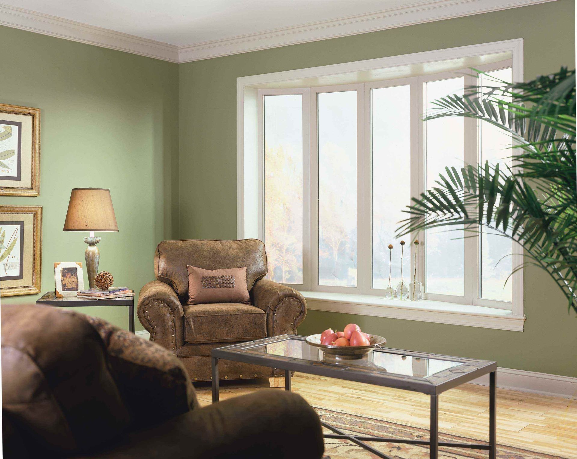 A cozy living room with sage green walls, a brown armchair, a glass coffee table, and a large bay window with a plant.