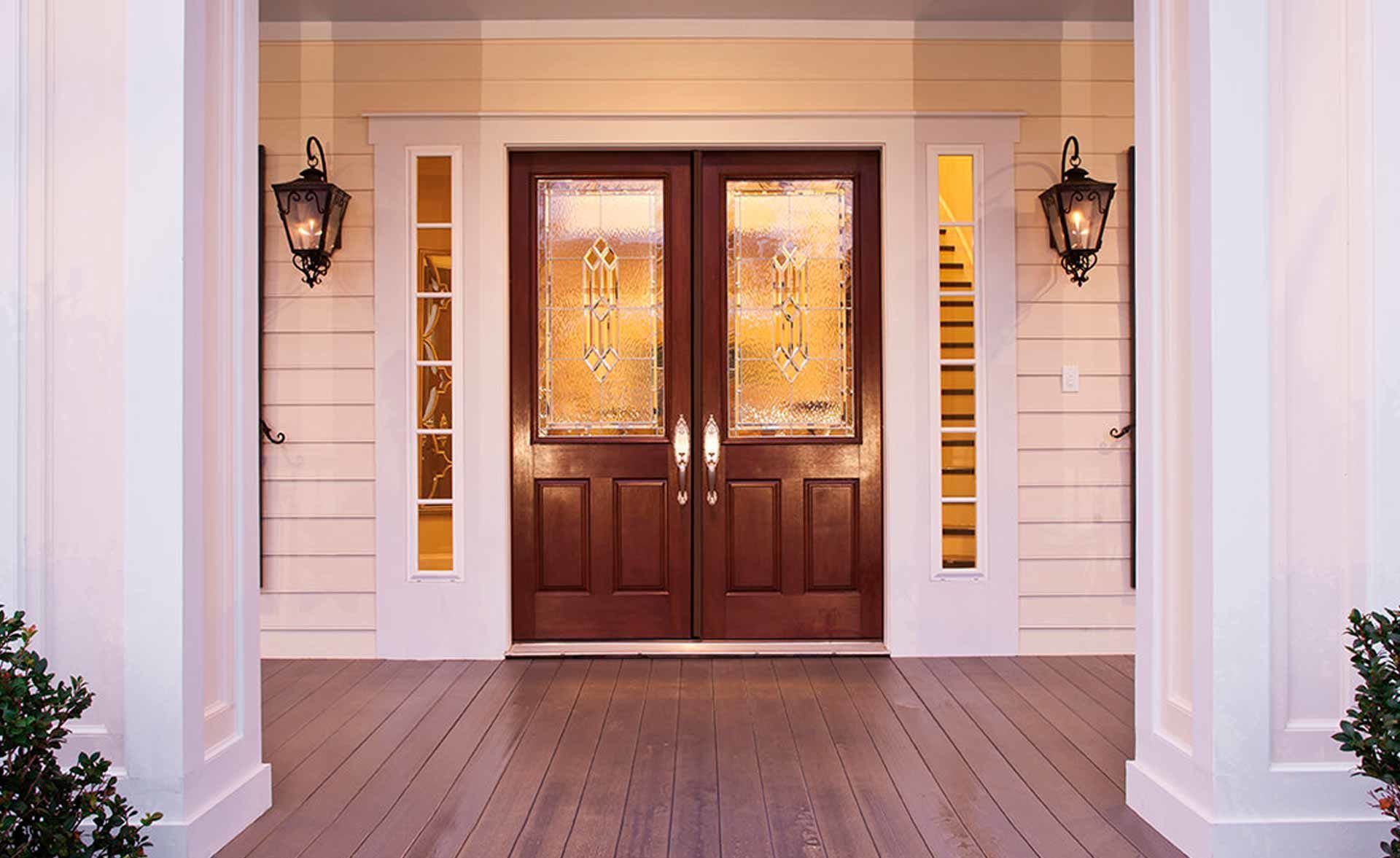 A front porch featuring a dark wood double door with glass panels, flanked by two wall-mounted lanterns and white columns.