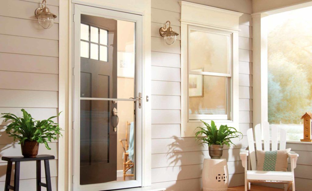 A bright porch with a white storm door, two potted ferns, a small stool, and a white chair under warm sunlight.