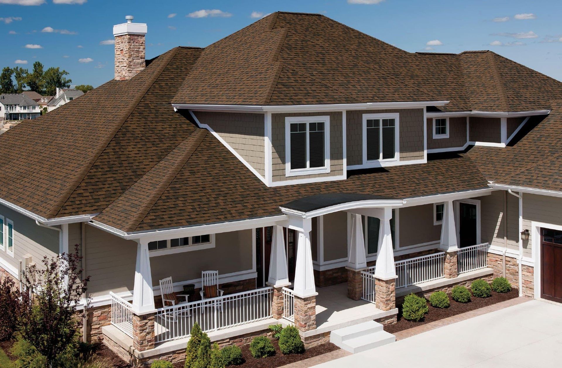 A suburban house with a tan exterior, stone foundation, and a dark brown shingled roof, featuring a covered front porch.
