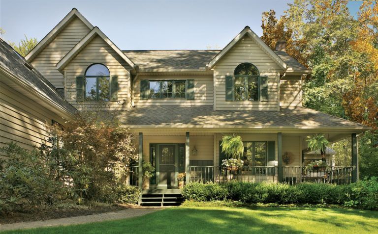 A two-story, tan-sided house with a front porch, arched windows, and a lush, tree-filled lawn.