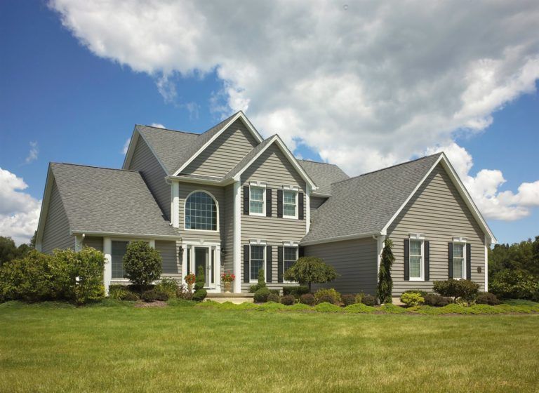 A two-story grey vinyl-sided house with a dark shingled roof, set against a blue sky with white clouds over a green lawn.