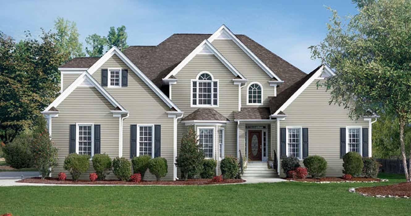 A two-story, beige-sided suburban house with dark gray shutters, a gabled roof, and a green lawn under a clear blue sky.