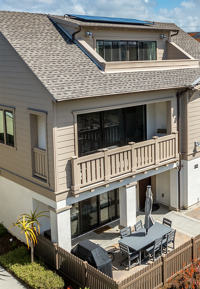 A tan two-story townhouse with a balcony, an upper-level dormer, and a ground-floor patio with a table and grill.