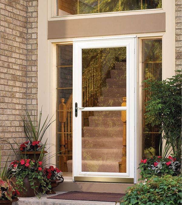 A white storm door with a large glass panel and a gold-tone kickplate installed in a brick entryway with potted flowers.