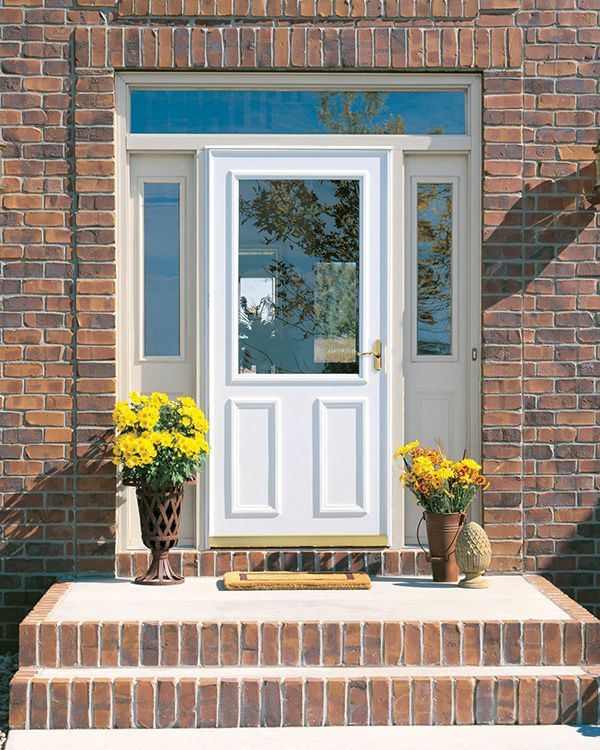 A white front door with a glass pane, flanked by sidelights and a transom, set in a brick exterior with two step planters.