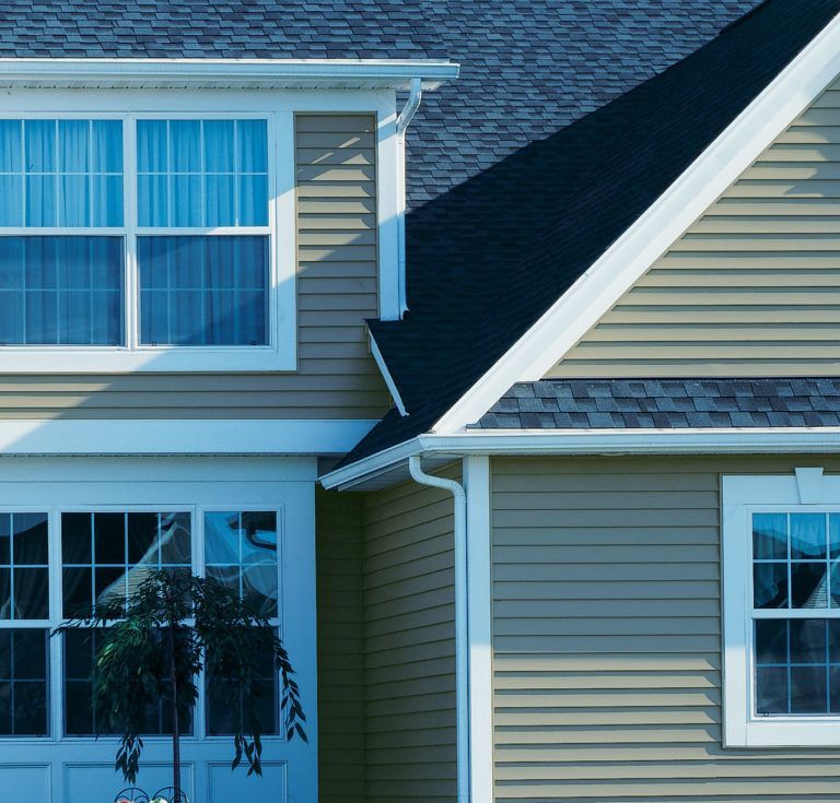 A view of a tan-sided house with a dark shingled roof, showing multi-pane windows and white trim.