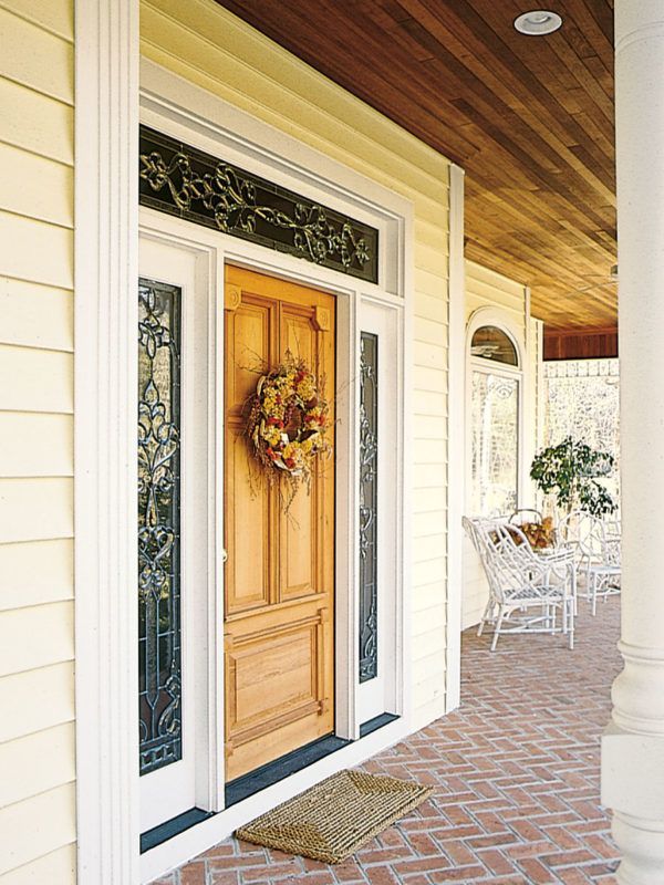 A natural wood front door with a decorative wreath, side glass panels, and a transom window on a porch with brick flooring.