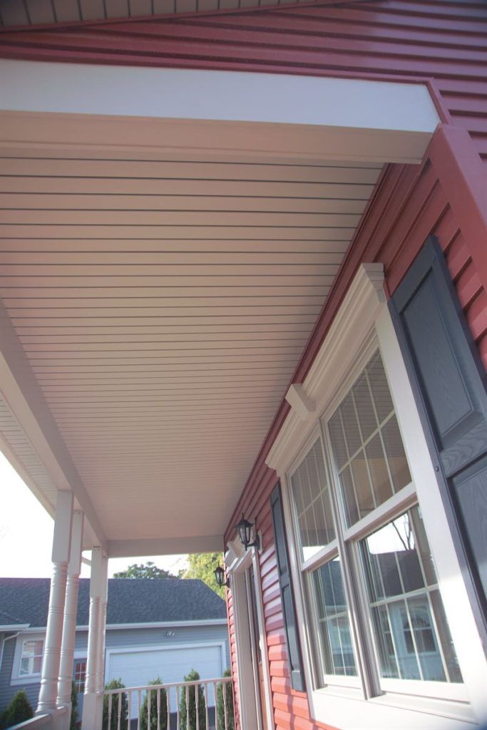 A view looking up at a covered front porch featuring a white slatted ceiling, red siding, and a window with black shutters.