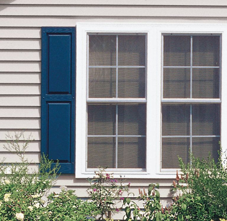 A white-framed window with blue shutters on light siding, partially obscured by green garden plants.