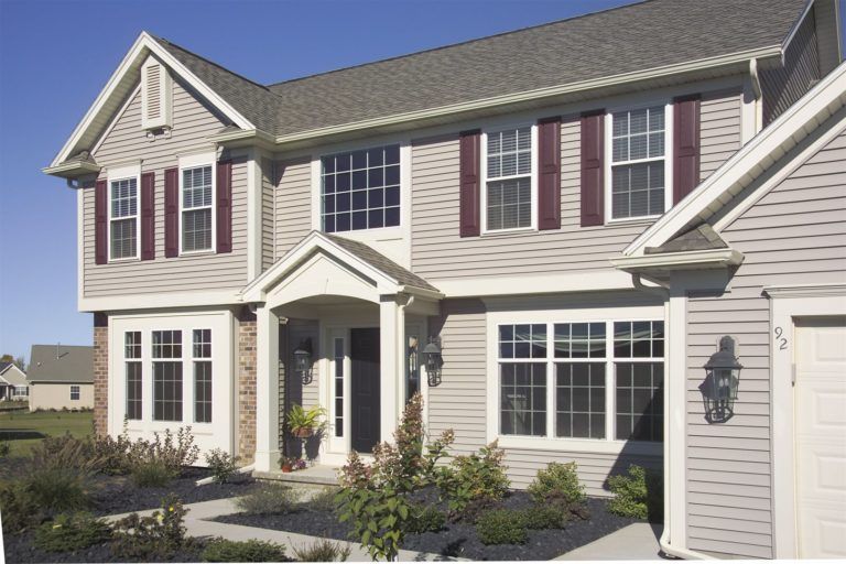 A two-story suburban house with light gray siding, dark shutters, a front porch, and a gravel garden bed in front.