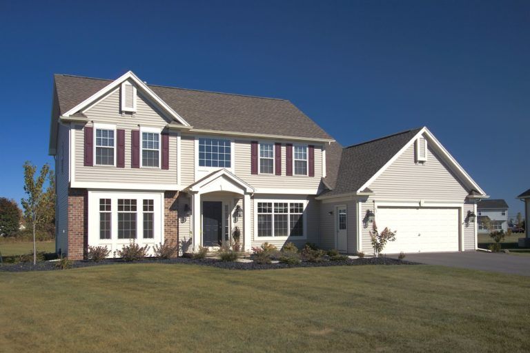 A two-story suburban house with beige siding, dark shutters, a front porch, and an attached two-car garage.