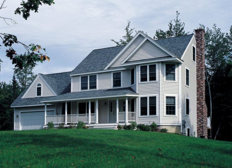 A two-story white house with a gray roof, front porch, attached garage, and a brick chimney, situated on a grassy lawn.