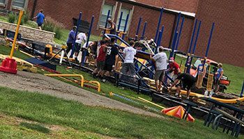 A group of people wearing casual clothing works together to assemble a playground structure on a grassy school field.