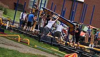 A group of people works together to lift and assemble a large, blue playground structure on a grassy school field.