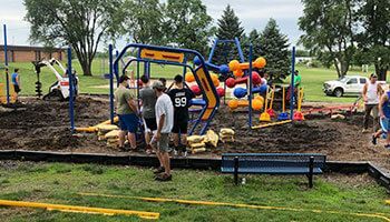 A group of people assemble a colorful modular playground structure on a construction site with equipment and trees nearby.