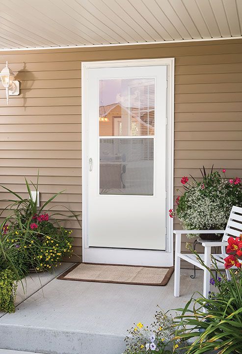 A white storm door with a large glass window pane set in a beige siding wall next to a porch with plants and a white chair.