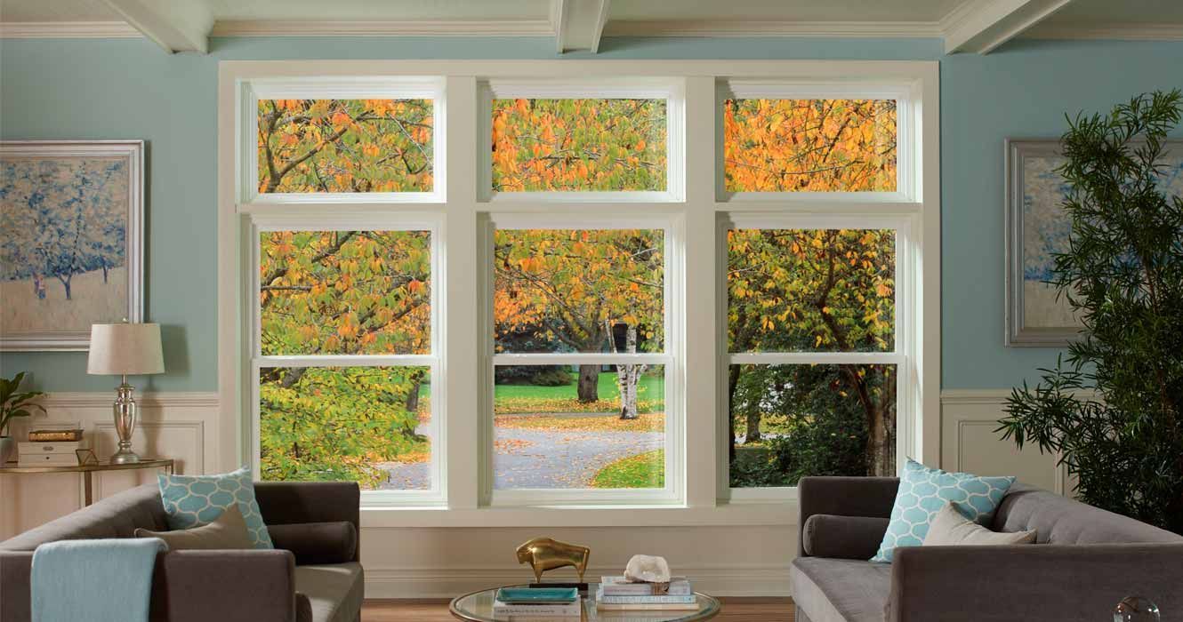 A bright, airy living room features a large triple window overlooking autumn trees, flanked by two sofas.