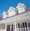 A light-colored house exterior with three gabled roof dormers, a porch, and dark blue window shutters against a blue sky.