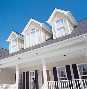 A light-colored house exterior with three gabled roof dormers, a porch, and dark blue window shutters against a blue sky.
