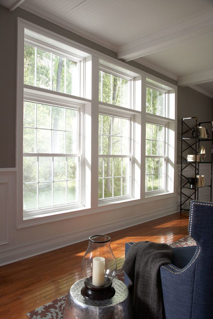 A brightly lit room with a wall of three large, white-framed windows looking out onto a wooded area.