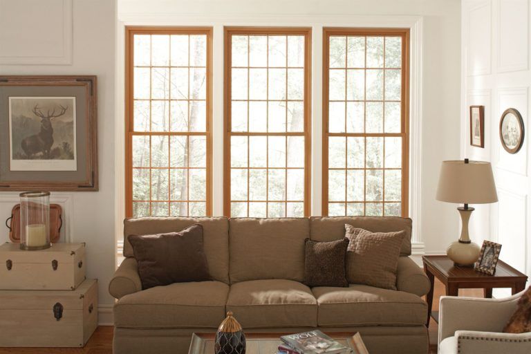 A cozy living room with a tan sofa in front of three tall, wood-framed windows, flanked by a side table and storage chests.