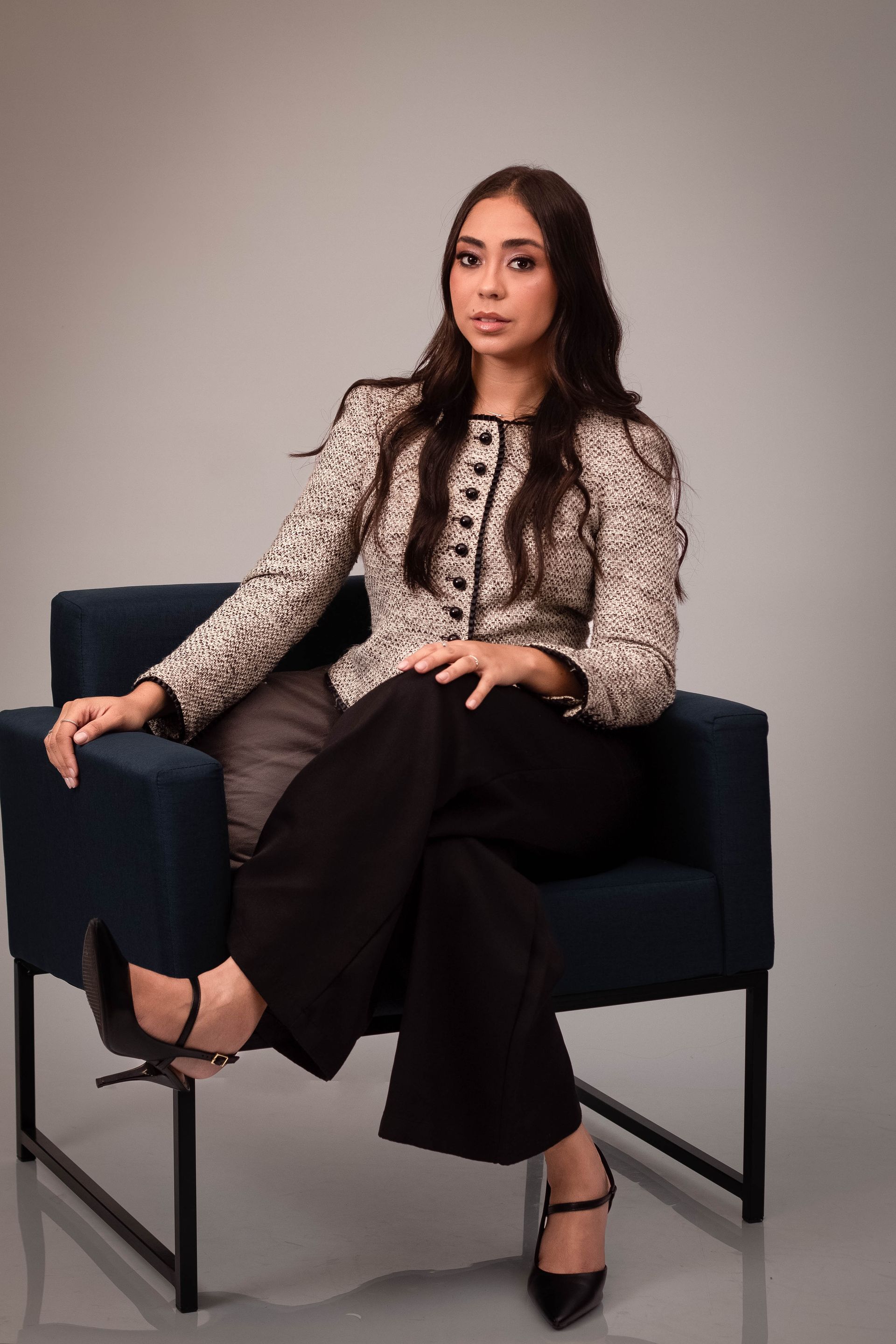 Mulher com jaqueta de tweed e calças pretas sentada em uma poltrona azul; pose angulada, expressão neutra.