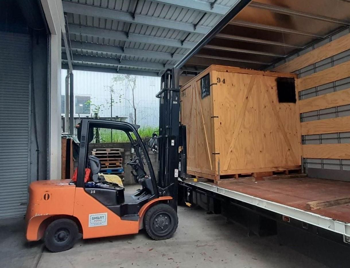 Orange Forklift Loading a Large Wooden Crate Onto a Truck in a Warehouse — John's Furniture Removals in Yandina, QLD