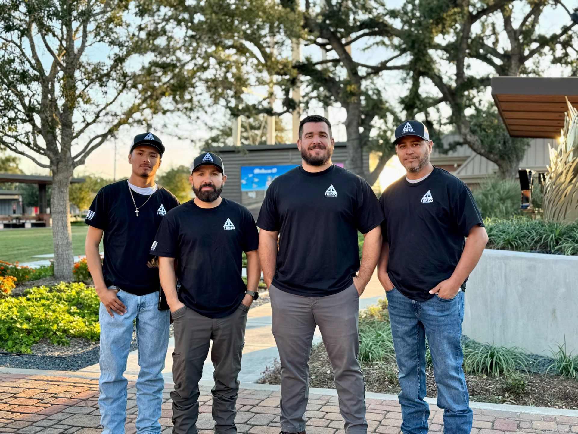 Four men in black shirts and hats stand outside; trees and a building in the background.