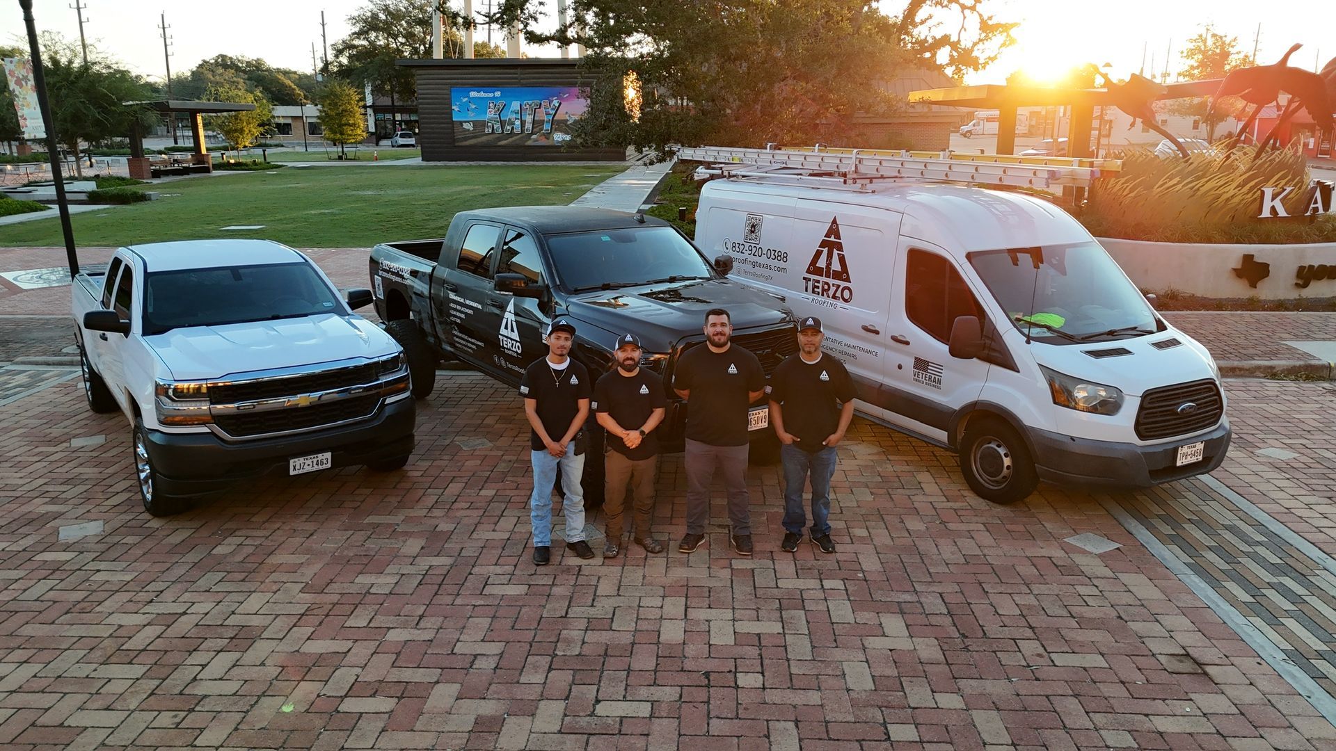 Four men stand in front of vehicles; a truck, a pickup, and a van. Evening sunlight. Brick pathway.