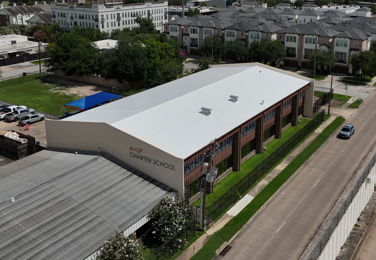 Aerial view of a long, light-colored building with a corrugated metal roof, next to a road with a car.